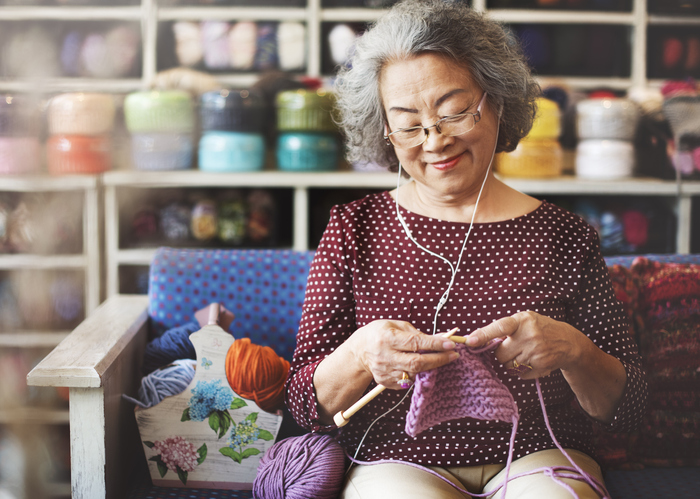 Elderly woman smiling while knitting and listening to music on in-ear headphones.